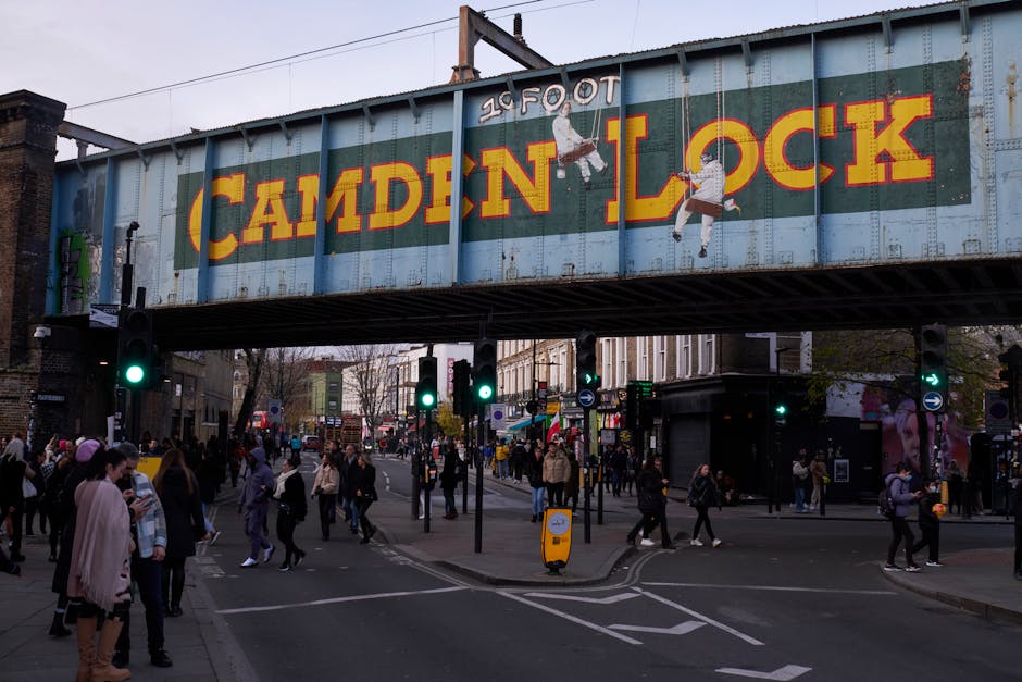 Wide view of Camden Lock railway bridge with a large painted sign reading 'Camden Lock' in yellow and red letters, spanning across a busy street in Camden, London. Below the bridge, numerous pedestrians are crossing the intersection, some waiting at traffic lights, while others are walking along the pavement. Several traffic signals display green lights, guiding the flow of people and vehicles. The scene includes a variety of shops and street vendors lining the sidewalk, with people carrying bags and engaging in commerce. The environment appears active, with a mix of daytime natural lighting and street illumination, consistent with urban busy district surroundings. The area reflects elements related to home or commercial relocation activities, with potential signage or equipment associated with moving or transport services, such as carts or packing supplies, although primarily features the urban transit scene. Man and a Van Camden occasionally operate in such environments, supporting furniture transport and packing logistics for local moves.