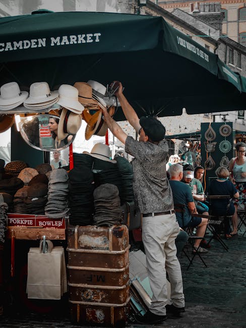 A man standing in front of a market stall under a green canopy labeled 'Camden Market' is reaching up to adjust hats displayed on the canopy's edge. The stall showcases a variety of hats, including straw hats, caps, and possibly fedoras, organized in stacks and hanging arrangements. A mirror reflecting part of the market is attached to the canopy, and a vintage brown suitcase with visible wear is placed on the ground nearby, along with a beige bag and some boxes. In the background, several market visitors are seated on chairs, some engaged in conversation, and attractions of the outdoor market can be seen, with market booths, decorative items, and other merchandise visible. The scene captures a lively outdoor environment typical of home relocation or packing and moving preparations, with the market setting suggesting careful packaging and transportation of items, supported naturally by the presence of packing materials and equipment.