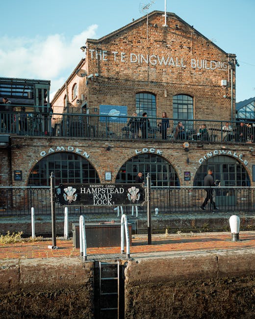Wide view of Camden Lock railway bridge with a large painted sign reading 'Camden Lock' in yellow and red letters, spanning across a busy street in Camden, London. Below the bridge, numerous pedestrians are crossing the intersection, some waiting at traffic lights, while others are walking along the pavement. Several traffic signals display green lights, guiding the flow of people and vehicles. The scene includes a variety of shops and street vendors lining the sidewalk, with people carrying bags and engaging in commerce. The environment appears active, with a mix of daytime natural lighting and street illumination, consistent with urban busy district surroundings. The area reflects elements related to home or commercial relocation activities, with potential signage or equipment associated with moving or transport services, such as carts or packing supplies, although primarily features the urban transit scene. Man and a Van Camden occasionally operate in such environments, supporting furniture transport and packing logistics for local moves.