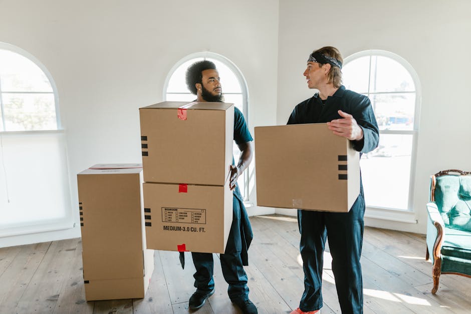 Two movers from Man and a Van Camden are outside a building, engaged in a home relocation process. They are carrying a large, green upholstered sofa with wooden legs, with one mover on each side suppo